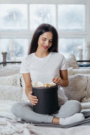 Happy young adult woman spending weekend at home, sitting on cozy bed with present in hands, smiling...