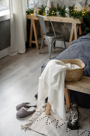 Vertical view of wicker basket with plaid on wooden bench near slippers and comfortable bed at cozy...