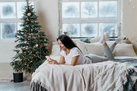 Side view of young adult girl spending christmas holidays at home, lying on bed, using smartphone, smiling wide and resting in bedroom with decorated new year treeの写真素材