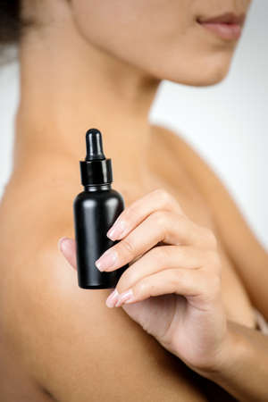 Concept of beauty treatment procedure. Vertical selective focus on glass bottle with anti aging organic serum oil. Young afro american woman holding cosmetics on white backgroundの写真素材