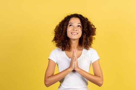 Please give me a sign. Happy and hopeful young african american woman with prayer hands looking up,...