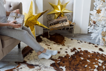 Cropped view of woman holding new year present in hands, sitting on comfort armchair in pajamas near...