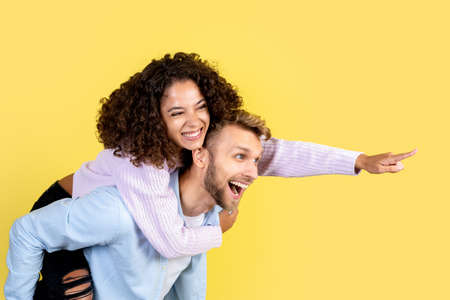 Happy european man carry on the back afro american woman while she pointing aside and smiling wide. People standing together on yellow background with copy spaceの写真素材