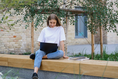 Remote education concept. Full length view of adult academy student girl smiling, using modern laptop computer, sitting near campus building and preparing homework for online coursesの写真素材
