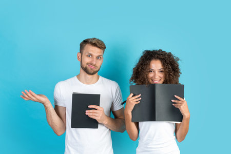 Education concept. Mixed race friends with hometask in exercise books standing together on blue background with copy space. Student making happy and sad faces, looking at cameraの写真素材