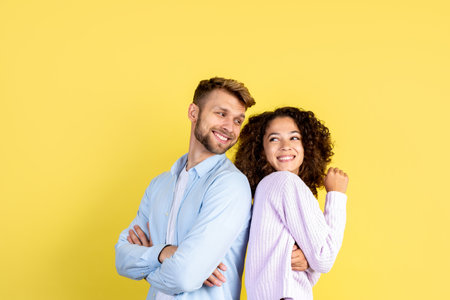 Two happy mixed race people standing back to back on yellow background with copy space, looking at each other, laughing wide and spending free time togetherの写真素材