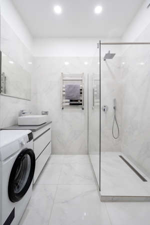 Apartment interior design concept. Vertical view of white modern bathroom with automatic washing machine, glass wall in shower cabin, mirror over sink and towel on heated rail at ceramic tiled wallの写真素材