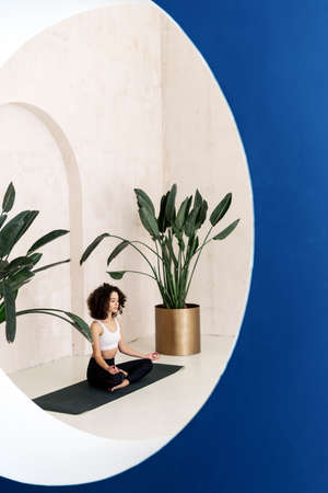 Mental health concept. Vertical view of calm african american woman sitting in meditation pose on exercise mat, spending time in yoga center. Focused lady making harmony and balance practiceの写真素材