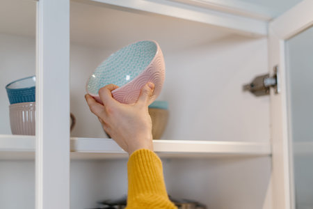 Cropped view of woman standing near open door at kitchen cabinet, puts clean ceramic dishes in the...