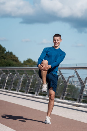 Concept of wellness and healthy lifestyle. Full length vertical view of happy young athletic guy stretching legs before running, holding balance, making sport training outdoors alone, doing exerciseの写真素材