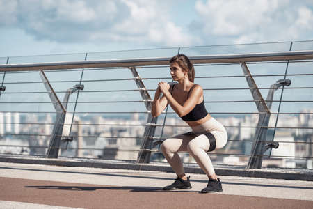 Morning workout concept. Focused adult athlete woman making sport training outdoors alone, squat with resistance band equipment, doing exercise on streetの写真素材