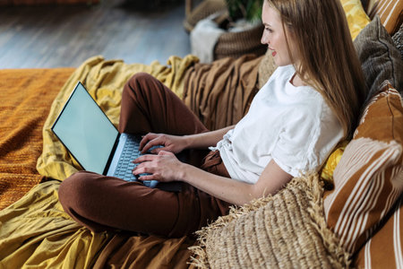 Profile side view of happy young adult woman using modern laptop computer with copy space on display, resting in bedroom, spending morning at home, sitting on bedの写真素材