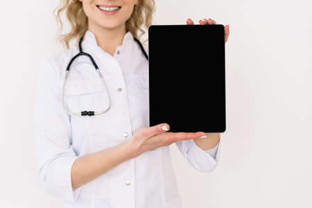 Cropped view happy family doctor in white medical uniform showing tablet computer with copy space display at camera. Nurse with empty blank screen on digital device remind about consultation onlineの写真素材