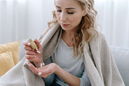 Sick woman covered with blanket holding capsule pills in hand. Concept of medicine for immunity during the global coronavirus covid-19 pandemic and quarantine. Health care and treatmentの写真素材