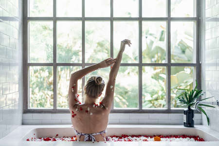 Body care concept. Back view of carefree young adult girl relaxing in bath with tropical flowers, raised hands up, spending morning in bathroomの写真素材