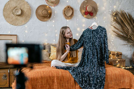 Happy young adult vlogger showing new trendy dress, creating media content for internet blog, recording video at broadcasting equipment, spending free time at homeの写真素材