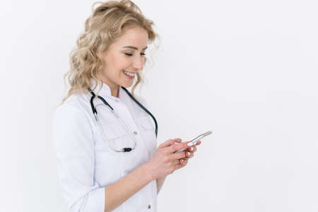 Mobile app and modern tech concept. Happy woman doctor in white medical uniform hold smartphone, reading message from patient, send online consultation standing isolated on copy space backgroundの写真素材