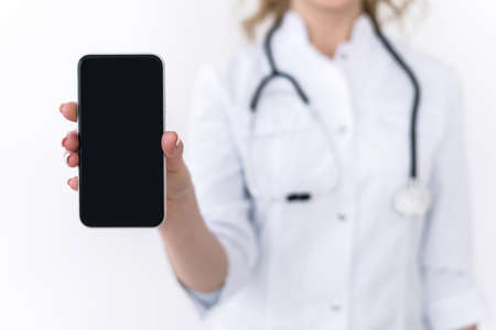 Cropped view of family doctor in white medical uniform hold modern smartphone with empty blank screen and copy space for advertising product or service. Woman holding modern gadget with mockup displayの写真素材