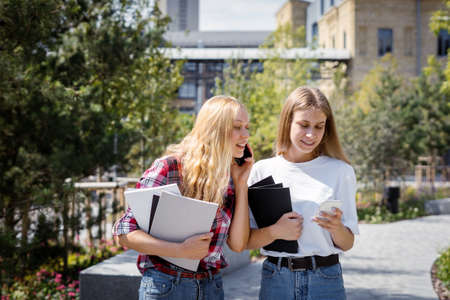 Two friends spending time near campus, using modern smartphone and walking outdoors with copybooks. Smiling adult academy student girl study together after classes and lecturesの写真素材
