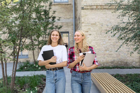 Two happy and smiling young academy student girl walking outdoors after study, holding copybooks in hands, spending time near campus together, smiling wide, talking to each otherの写真素材