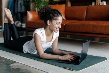 Happy young african american woman using modern laptop computer, lying on exercise mat in...