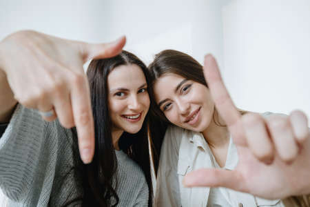 Portrait of best friends looking at camera, smiling, standing in living room at home. Two women making a frame gesture with their fingers in front of faces as visualises an idea or compositionの写真素材