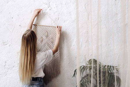 Back view of young adult woman hangs white handmade macrame on the wall in room with cozy interior....