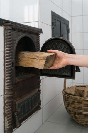 Cooking with minimum environmental impact. Vertical shot of male hand putting firewood in opened firebox of wood burning stove made of glazed white tiles. Traditional home heating conceptの写真素材