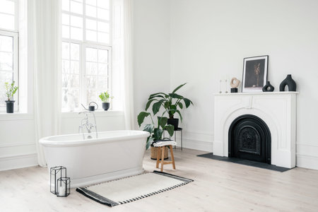 White classic freestanding bathtub, large windows, modern fireplace and green houseplants in light...