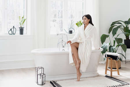 Body care, skincare and natural beauty concept. Full length view of young asian woman sitting on freestanding bath in white bathrobe, looking a side. Beautiful female in modern interior bathroomの写真素材