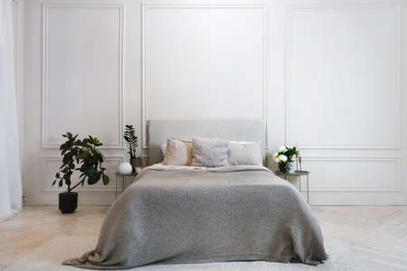 Large comfortable bed with gray cover and comfy pillows in spacious stylish modern white bedroom in minimalistic design with green plants and flowersの写真素材