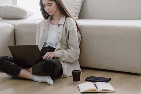 Online education concept. Full length view young concentrated student make homework. Woman working at home office with laptop, sitting on floor near comfortable sofa in living room, wearing in casualの写真素材