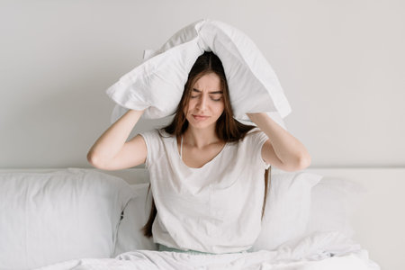 Upset and tired young woman sitting on bed in pajamas, covers ears with a pillow, drowns out the...