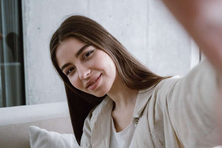 Blogger recording video or making online call. Portrait view of young millennial woman standing in living room, take self portrait for social networks on camera, smiling wide. Lifestyle conceptの写真素材