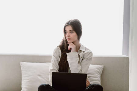 Thoughtful young woman sitting with laptop on couch, looking aside, thinking about business strategy or startup project. Student in a beige shirt study at home, spending time in living roomの写真素材