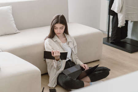 Full length view of young woman with laptop holding notepad in hands. Concentrated and smart student girl in casual wear sitting on floor near comfortable sofa in living room, make homework taskの写真素材
