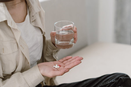 Cropped view of young unhealthy woman hold glass of water and treatment pills in her hand. Female take painkiller medication for throbbing headache, reducing, medical abortion or season allergyの写真素材