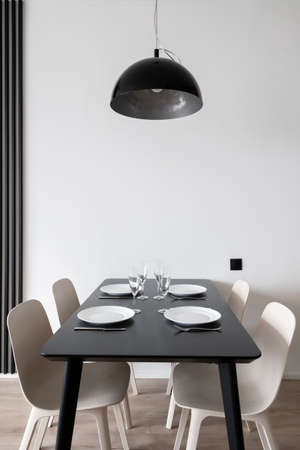 Vertical shot of large black kitchen table setting for family dinner with dinnerware, wineglasses and cutlery with hanging metal lamp above and white chairs in minimalistic interior designの写真素材