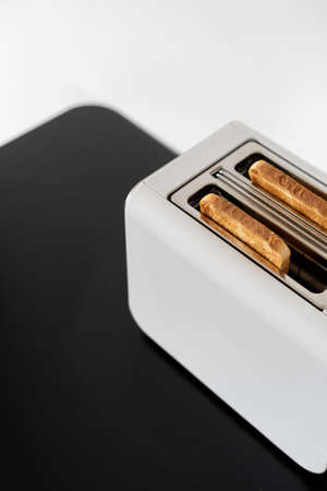 Close up of white toaster with delicious crispy slices of bread inside on black table against white wall, breakfast ready to eat. Modern kitchen cooking appliance conceptの写真素材