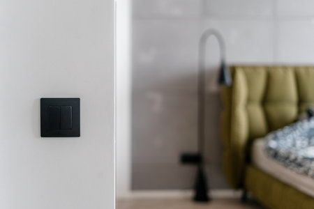 Close up of simple black matte light switch on white wall with bed and metal standing lamp in the...