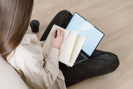 Rear view of young woman writing in notebook with copy space pages, sitting on floor in living room near blank screen laptop display. Female make home task notes in diary, education conceptの写真素材