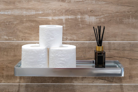 Modern metal chrome shelf with aroma sticks and rolls of toilet paper on wall with wood pattern structure on background. Minimalist interior design in bathroomの写真素材