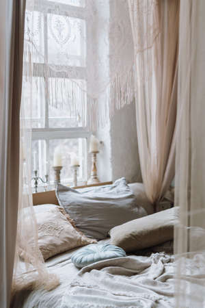 Vertical shot of cozy bed with various pillows and open baldachin, with light coming from big white vintage window with many different candles on windowsill. Empty bedroom in morningの写真素材