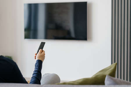 Modern living room interior with blank tv screen panel on blurred white wall background. Cropped shot of woman with remote control in hand sitting on sofa in room with minimalist functional designの写真素材