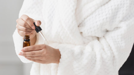 Cropped shot of female model in white bathrobe applying beauty drops taking them from glass bottle, putting on hand using pipette during morning beauty routine at bathroom, testing organic cosmeticの写真素材