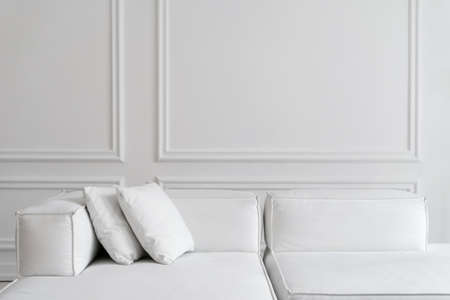 Stylish contemporary designer white sofa with two cushions in room with white walls and high ceiling, sun rays shining through window, cropped shot. Minimalistic design conceptの写真素材