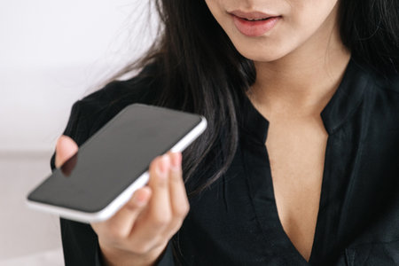 Cropped shot of young woman recording voice message on mobile phone with mockup display, focus on person. Female calling and getting in touch with friends and family while spending day at homeの写真素材