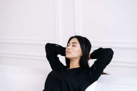Carefree young lady relaxing on couch indoors, dreaming with closed eyes and enjoying leisure time alone at home. Beautiful sleepy asian woman having rest and taking nap in light modern living roomの写真素材
