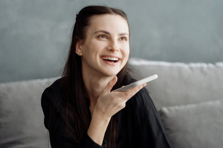 Distant communication concept. Young happy woman recording voice message on smartphone. Smiling...
