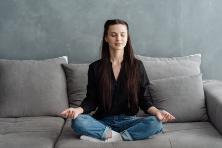 Relaxed serene young woman meditating, sitting on comfort couch in living room, feel fatigue after...